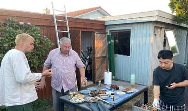 Drei Männer genießen ein Barbecue im Garten. Auf dem Tisch stehen jede Menge Grillgerichte und Getränke. Ein Schuppen, eine Leiter und üppiges Grün runden das Bild ab.