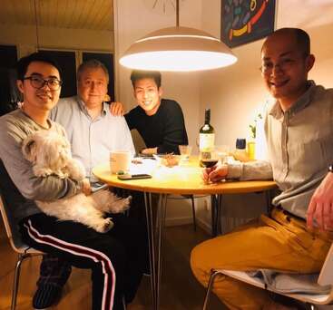 Vier Männer sitzen bei warmer Beleuchtung um einen Tisch herum. Einer hält einen flauschigen weißen Hund im Arm. Es gibt Wein, Snacks und freundliche Lächeln, was eine gemütliche, entspannte Atmosphäre schafft.