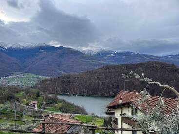 A tranquil landscape featuring red-roofed houses overlooking a lake, surrounded by green hills and forests, with distant snow-capped mountains beneath a dramatic cloudy sky.