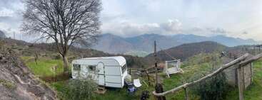 A white caravan sits on a grassy hillside, surrounded by mountains, a tall leafless tree, wooden swing, scattered chairs, rustic fence, under a cloudy sky.