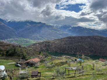 Rolling green hills overlook a calm river, with rustic houses and wooden fences. Snow-capped mountains tower in the background under a dramatic, cloud-filled sky. Peaceful scenery.