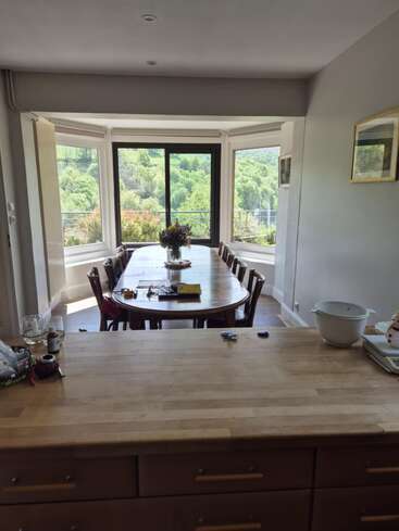 Une salle à manger spacieuse comprend une grande table en bois avec des chaises, un bouquet en centre de table, des fenêtres panoramiques offrant une vue sur une végétation luxuriante et un îlot de cuisine en bois à l'avant.