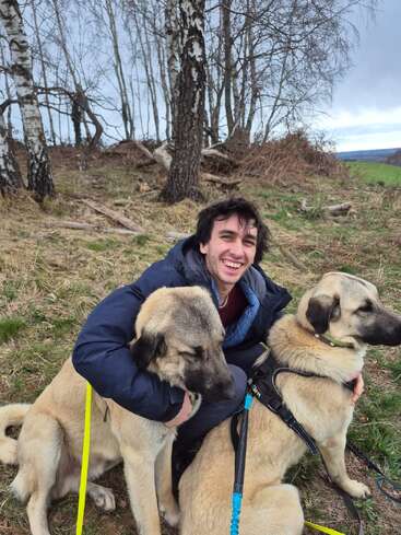 Une personne souriante est assise dehors, serrant dans ses bras deux grands chiens bruns tenus en laisse. Derrière eux se dressent des bouleaux dénudés, et le ciel est nuageux, laissant présager un temps frais.