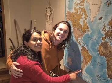 Un couple souriant se tient debout à l'intérieur, montrant du doigt une grande carte du monde accrochée au mur. L'atmosphère semble chaleureuse et conviviale, suggérant l'excitation du voyage.