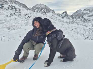 Une femme vêtue de vêtements d'hiver rit joyeusement tandis que son chien noir lui donne affectueusement des petits coups de museau. Ils sont entourés de montagnes enneigées sous un ciel nuageux.