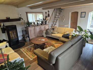 Ce salon confortable comprend deux canapés, un poêle à bois, du parquet, des plantes, une table ronde, des tapis décoratifs, un meuble, des escaliers et des décorations murales.