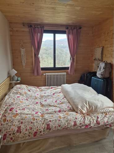 Cette chambre confortable en bois est dotée d'un couvre-lit à fleurs, de rideaux roses, d'une grande fenêtre offrant une vue panoramique, de bagages et d'une décoration bohème qui lui confèrent une atmosphère paisible et rustique.