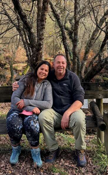 Um casal feliz está sentado bem juntinho num banco de madeira na floresta. Estão vestidos com roupas quentes, sorrindo, rodeados por árvores sem folhas e folhas caídas.