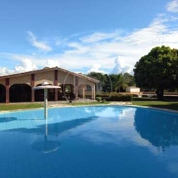Uma cena tranquila ao ar livre com uma grande piscina, uma fonte com um guarda-sol, um prédio de tijolos com arcos, árvores verdejantes e um céu azul brilhante.
