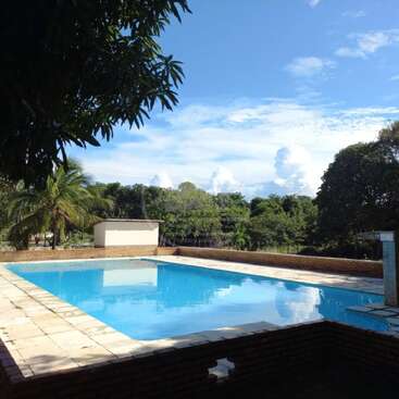 Uma piscina ao ar livre tranquila reflete o céu azul e as nuvens esparsas, cercada por árvores e um pátio de tijolos, com um pequeno prédio nas proximidades que abriga as instalações de serviço.