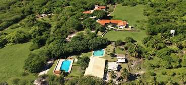 Esta imagem aérea mostra uma grande propriedade cercada por uma vegetação exuberante, com vários edifícios de telhado vermelho, duas piscinas, campos abertos e palmeiras espalhadas.