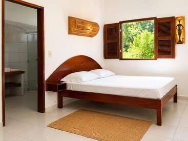 Este é um quarto luminoso e minimalista, com uma cama de madeira, roupa de cama branca, janela aberta, luz natural, quadros na parede, tapete tecido e um banheiro revestido de azulejos ao lado.