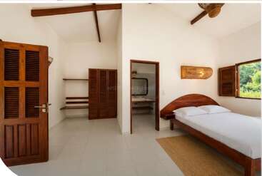 Esta imagem mostra um quarto claro e minimalista com móveis de madeira, uma cama de solteiro, paredes brancas, piso de azulejo, janela aberta, um armário e um banheiro anexo.