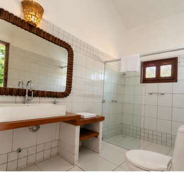 Esta casa de banho tem paredes e piso revestidos com azulejos brancos, um box de vidro, um espelho com moldura de madeira, um lavatório branco, um vaso sanitário, uma janela e uma iluminação suave.
