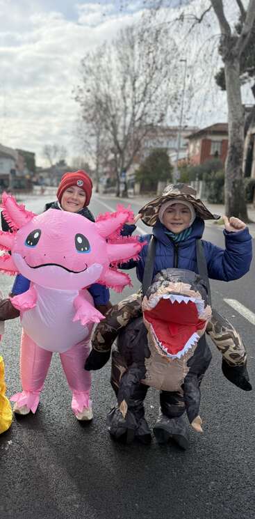Dos niños están de pie en una calle con divertidos disfraces hinchables: uno de axolotl rosa y otro de dinosaurio. Ambos sonríen, rodeados de árboles invernales y una carretera desierta.