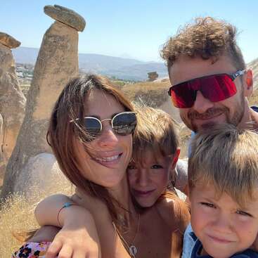 The image depicts a family of four posing for a selfie in a desert landscape, with large rock formations and mountains visible in the background on a sunny day.