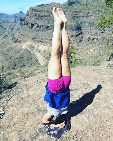 Uma mulher está de cabeça para baixo em um afloramento rochoso, com as pernas levantadas no ar, em uma paisagem montanhosa sob um céu azul.