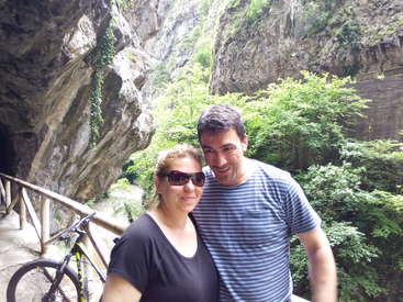 The image depicts a couple posing for a photo in a scenic outdoor setting, with a bicycle leaning against a railing behind them.
