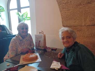 Two elderly women sit together at a restaurant table, smiling warmly. A cake and a gift bag are displayed, suggesting a celebration in a cozy setting.