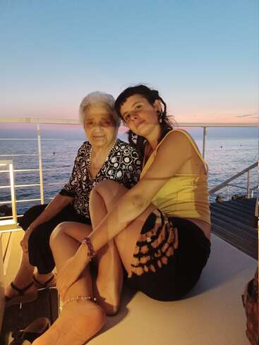 An elderly woman and a younger woman sit closely together on a deck by the sea at sunset, enjoying a peaceful moment and beautiful scenery.