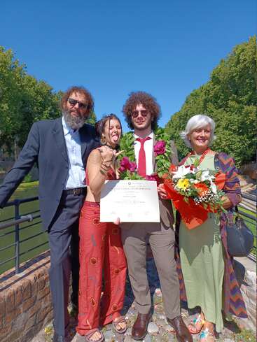A happy group celebrates a graduation outdoors. The graduate holds a diploma and wears a laurel wreath, while family members smile and pose with vibrant flowers.