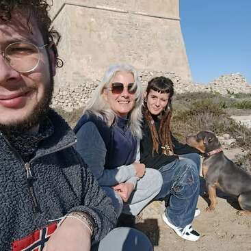 Three people and a dog sit together outdoors in the sunshine, smiling. They appear relaxed and happy, with an old stone building in the background.