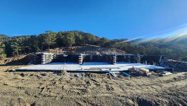 Eine Baustelle in einer ländlichen, hügeligen Gegend mit einem großen Betonfundament, Stützmauern, verstreuten Baumaterialien und Bäumen im Hintergrund unter einem klaren Himmel.