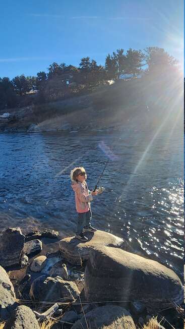 Un niño pequeño está de pie sobre unas grandes rocas junto a un río cristalino, pescando bajo la luz del sol, rodeado de naturaleza, con árboles y una casa al fondo.
