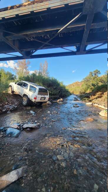 Un todoterreno blanco circulando por un camino de montaña junto a un arroyo rocoso bajo un puente metálico, rodeado de matorrales y árboles, con un cielo azul y despejado sobre él. Una escena de aventura en plena naturaleza.