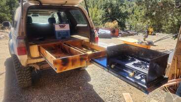 Un vehículo con el maletero abierto deja al descubierto unos cajones de madera hechos a medida y una cocina de camping extraíble con una cocina de dos fuegos, situada en un pintoresco entorno al aire libre.