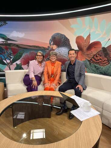 Tres personas están sentadas sonriendo en un sofá color crema en un estudio de televisión, con un vibrante mural de pájaros, flores y una mariposa al fondo.