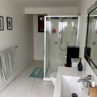 L'image représente une salle de bains aux murs et au sol blancs, avec une douche, un lavabo, des toilettes, un miroir et une œuvre d'art, avec un filigrane subtil au centre.