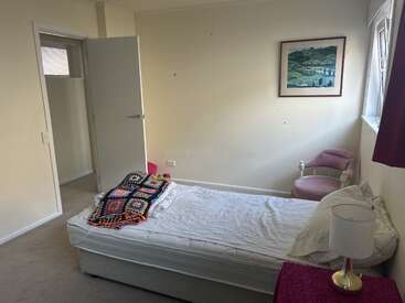 Dieses gemütliche Schlafzimmer verfügt über ein Einzelbett mit einer bunten Decke, einen rosa Stuhl, ein Landschaftsgemälde, eine Lampe, eine offene Tür und Tageslicht, das durch die Fenster hereinströmt.