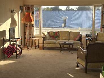 Ein gemütliches Wohnzimmer mit Vintage-Möbeln, großen Fenstern, geblümten Kissen, einem Retro-Lampenschirm, hereinströmendem Sonnenlicht, einem Fernseher auf einem Ständer und Teppichboden.