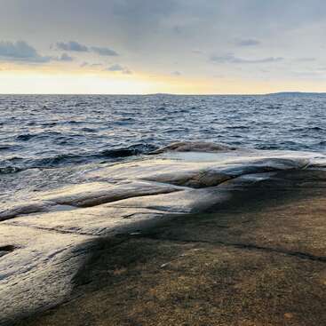 The image depicts a serene rocky shoreline, with a vast expanse of dark blue water stretching out to meet a cloudy sky at sunset.
