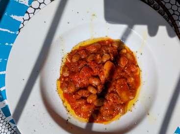 Une assiette blanche contient une portion de haricots dans une riche sauce tomate, arrosée d'huile d'olive. Les ombres et la lumière du soleil accentuent l'aspect vibrant et texturé du plat.