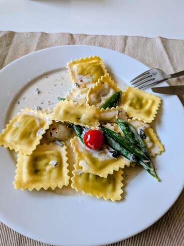 Une assiette blanche contient des raviolis dans une sauce crémeuse, garnis d'une tomate cerise et de haricots verts. Une fourchette et un couteau en argent reposent sur le bord de l'assiette.
