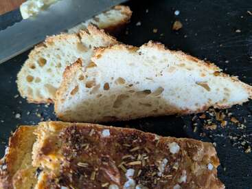 Cette image montre deux tranches de pain artisanal rustique à la mie aérée, l'une garnie d'herbes et de sel de mer, à côté d'un couteau à pain dentelé.