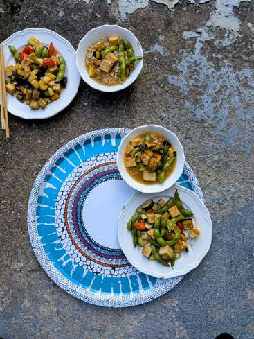 Quatre assiettes et bols de légumes colorés sautés avec du tofu et de l'edamame, certains avec des nouilles, sont disposés sur un plateau à motifs et une surface en béton usé par les intempéries.