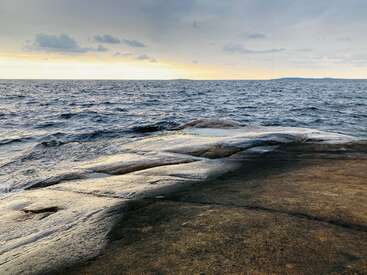 L'image représente un rivage rocheux serein, avec une vaste étendue d'eau bleu foncé qui s'étend à la rencontre d'un ciel nuageux au coucher du soleil.