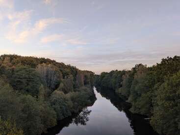 Une rivière calme serpente à travers une forêt dense et verte sous un vaste ciel pastel. L'eau reflète les arbres et les nuages, créant un paysage paisible et serein.