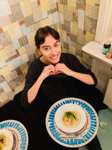 A smiling person sits at a table with a colorful patchwork wall behind them. They form a heart shape with their hands, enjoying a plate of pasta.