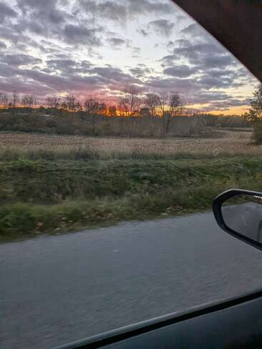Una vista desde la ventanilla de un coche muestra una carretera rural, campos y árboles desnudos con un dramático cielo nublado al atardecer que brilla en naranja y púrpura en el horizonte. Una escena apacible.