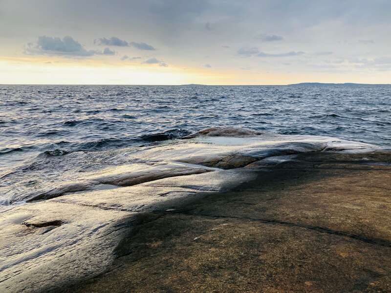 L'image représente un rivage rocheux serein, avec une vaste étendue d'eau bleu foncé qui s'étend à la rencontre d'un ciel nuageux au coucher du soleil.