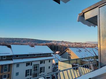 Several buildings with snow-covered rooftops face a scenic view of distant hills and a forest under a clear blue sky, bathed in morning sunlight.
