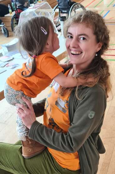 A smiling woman enthusiastically holds a young girl. Both wear orange shirts, sharing a joyful moment indoors, surrounded by colorful lines, papers, and wheelchairs in the background.