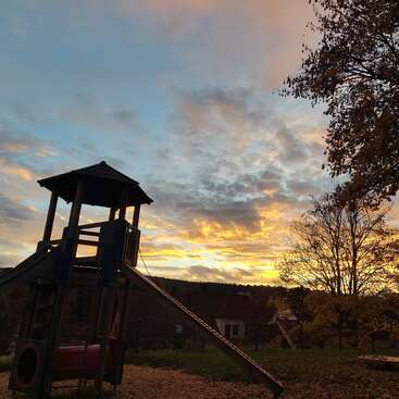 The image shows a playground tower with a slide at sunset. Vibrant clouds fill the sky, casting beautiful light. Trees and houses are visible in the background.