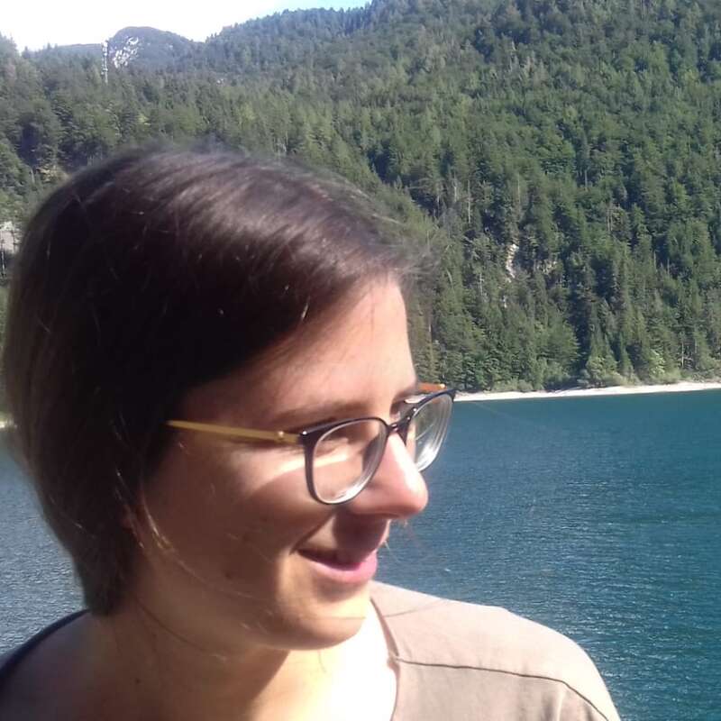 The image depicts a woman with short brown hair and glasses standing in front of a serene lake and forested mountain backdrop on a sunny day.