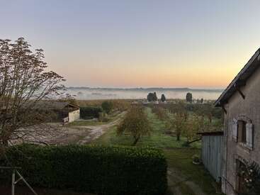 Un apacible paisaje rural al amanecer presenta bruma sobre los campos, árboles con hojas otoñales, rústicos edificios agrícolas y un cielo despejado con colores cálidos en el horizonte.