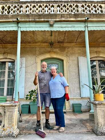 Una pareja sonriente posa junta en un porche rústico, sosteniendo una escoba. Parecen felices y contentos, rodeados de macetas y de una arquitectura antigua y encantadora.